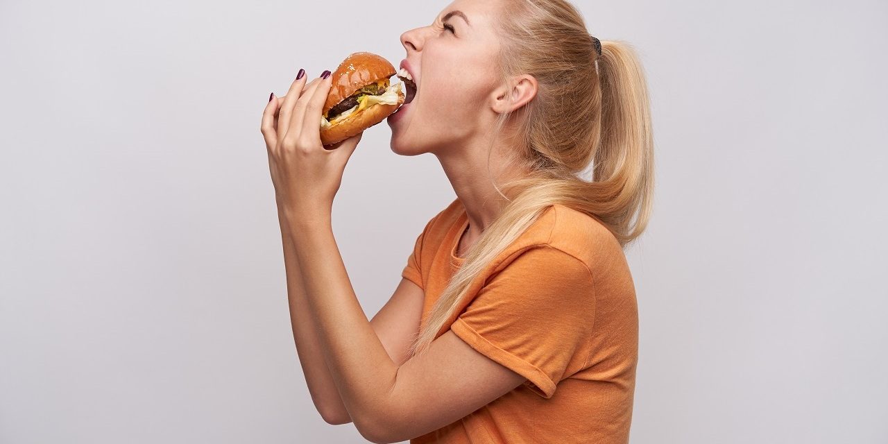 Back view of hungry young long haired blonde lady with casual hairstyle eating greedily big fresh burger while standing against white background and keeping her eyes closed