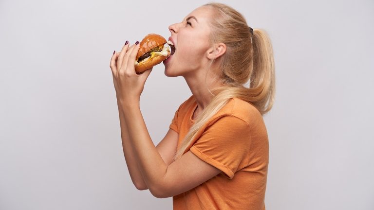 Back view of hungry young long haired blonde lady with casual hairstyle eating greedily big fresh burger while standing against white background and keeping her eyes closed